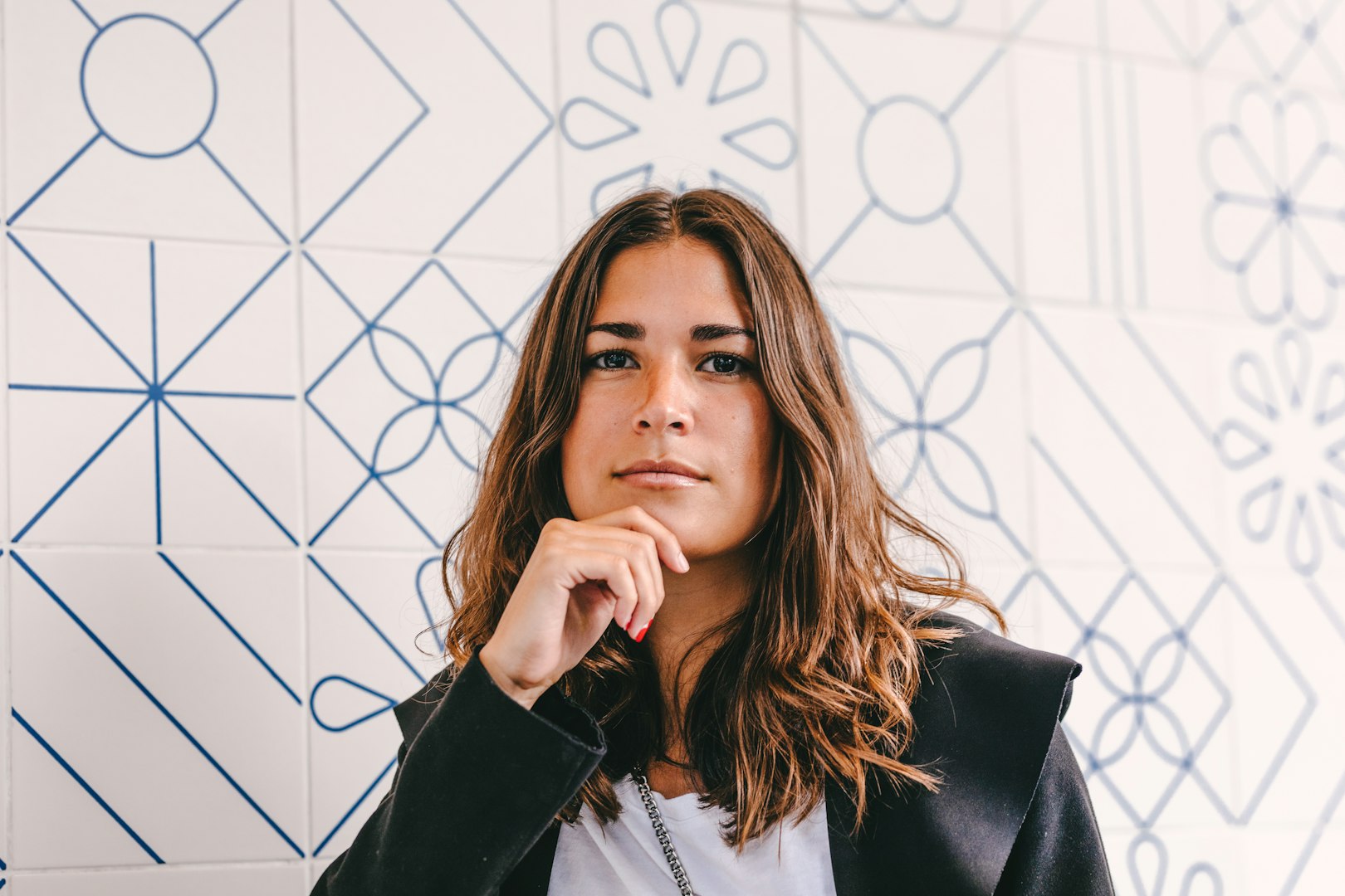 A woman with wavy, shoulder-length brown hair is smiling gently. She is wearing a white blouse and is standing in a bright, minimalist setting with turquoise decorative items in the background.